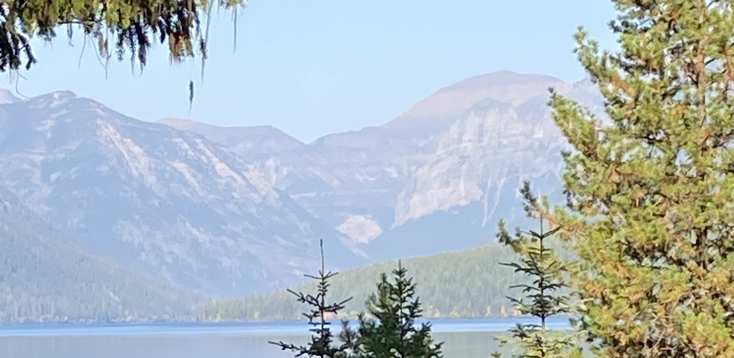 Beautiful rugged mountain scene with pine covered foothills and a glass smooth lake at base of mountains with pine trees in the forground