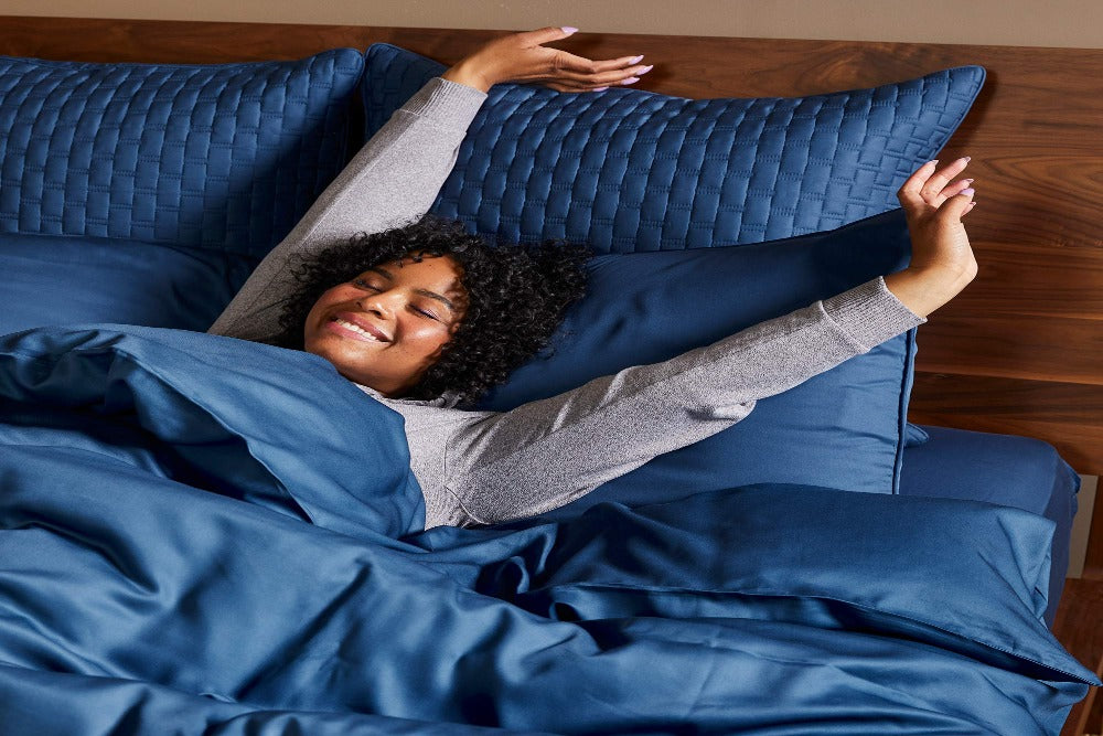 Happy attractive woman lying in bed stretching her arms and covered with a Luxurious 100% Bamboo Duvet Cover and four pillows at the headboard with matching 100% Bamboo Shams 