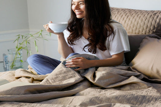An attractive happy woman holding a mug containing a hot beverage and sitting cross legged on a bed with her legs partially covered with a Champagne Colored Luxurious 100% Bamboo Quilted Coverlet