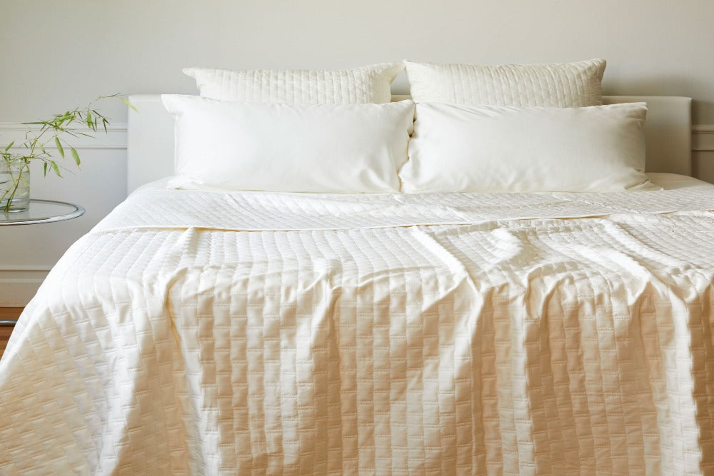  View of a loosely made bed in a light colored well decorated bedroom with an Ivory Colored Luxurious 100% Bamboo Quilted Coverlet draped over the bed and four matching pillows at the headboard