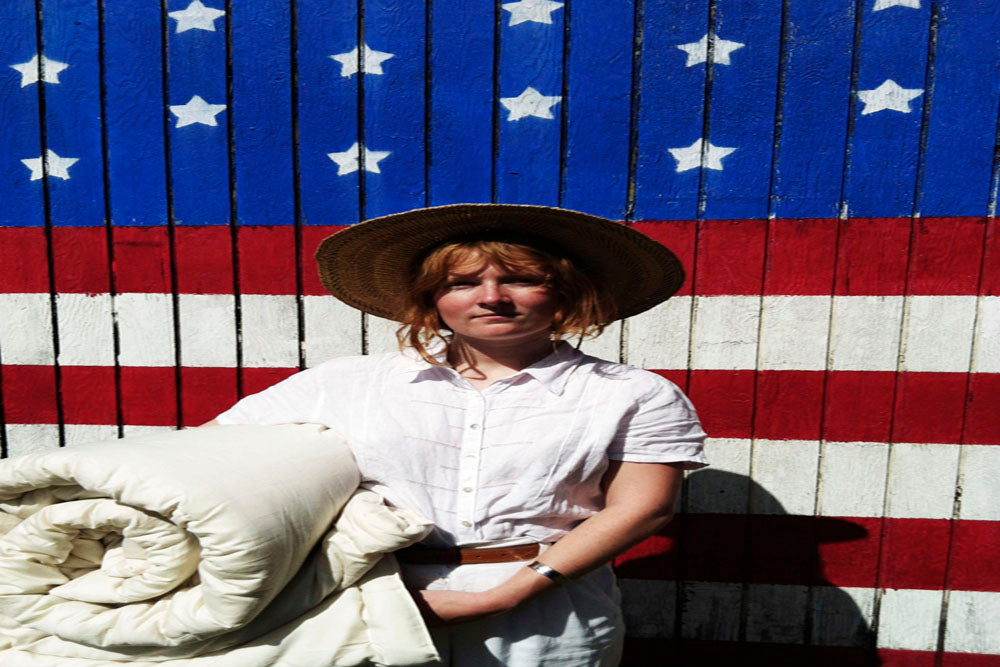 an attractive woman wearing a summer straw hat and dressed in organic cotton shirt and pants holding a loosely rolled up natural color certified organic comforter and standing in front of an American flag mural