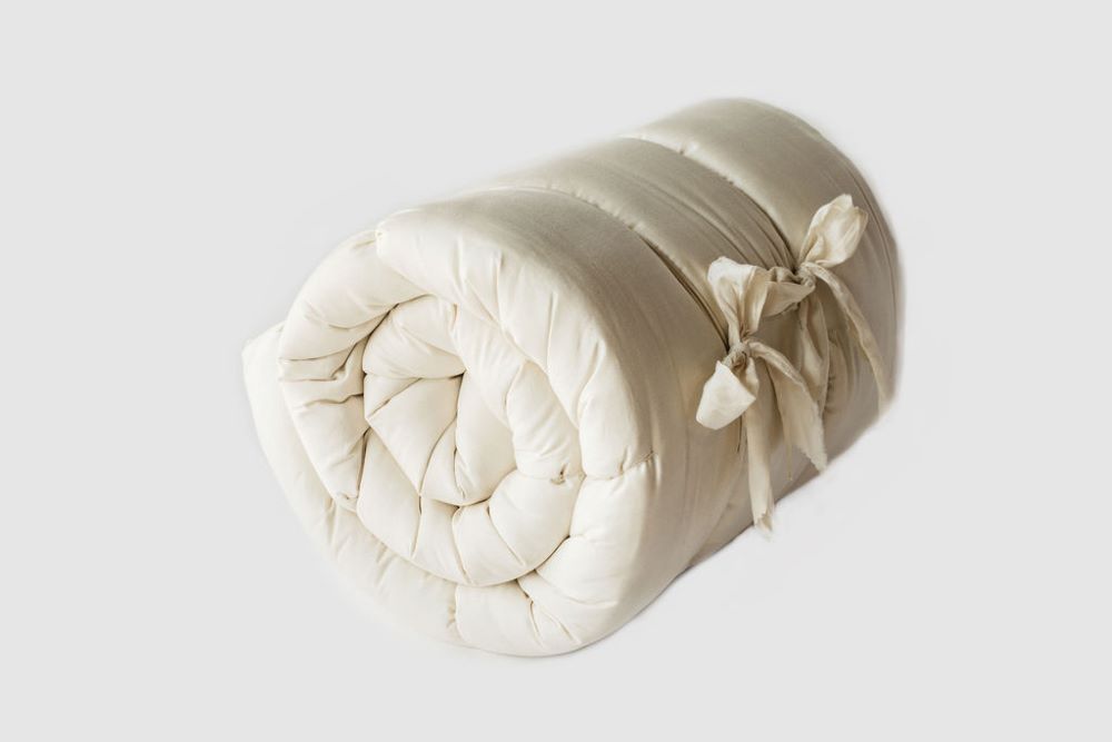 A quilted natural color organic mattress topper rolled up and tied with two cotton strips with each strip tied into a bow