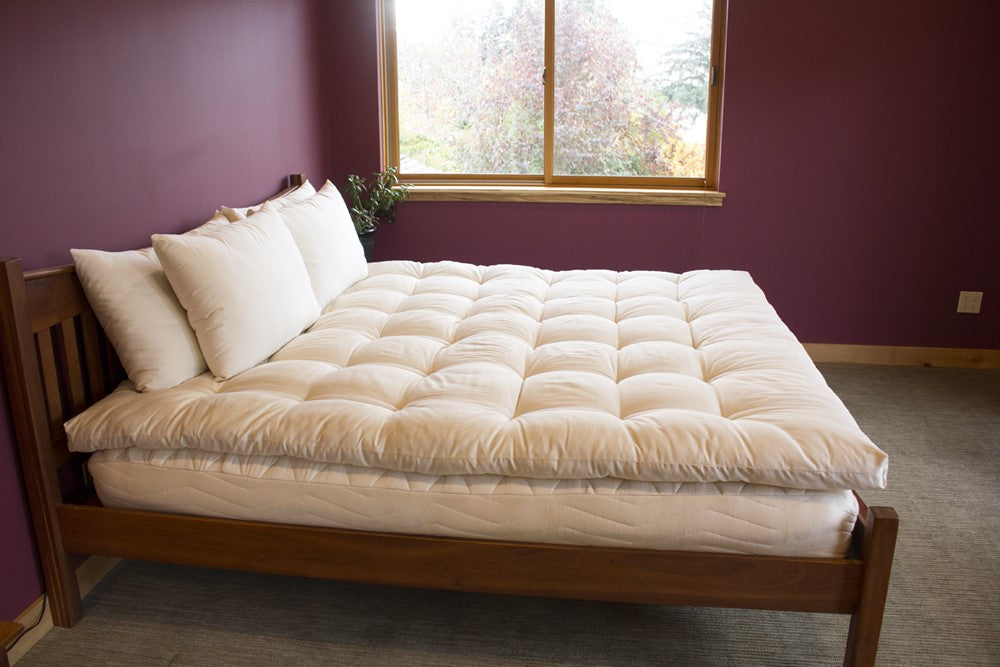 Side view of a wood frame unmade bed displaying a quilted cotton sateen mattress topper stuffed with eco wool and four organic cotton pillows at the headboard